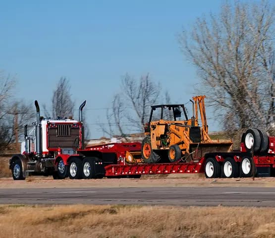 Tractor pulling a red lowboy trailer loaded with a yellow backhoe loader on a highway